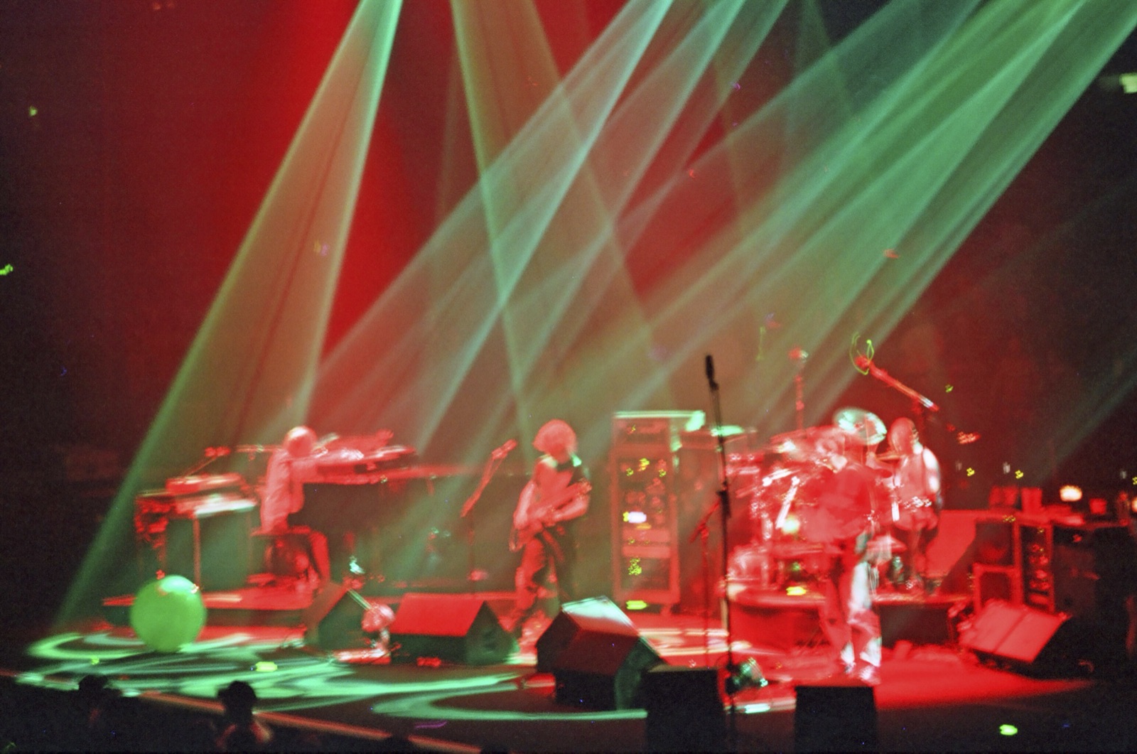 A band plays keys, guitar and drums on a stage flooded red and crossed by green light beams.